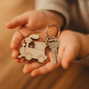 Può includere: Un portachiavi a forma di treno in legno con una chiave d'argento attaccata. Il portachiavi è tenuto in mani aperte. Il treno ha un design dettagliato con ruote, una finestra e fumo. La chiave è d'argento e ha una forma standard.