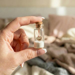 Peut inclure: Petite bouteille en verre transparent avec bouchon en liège, contenant du sable blanc et un minuscule cœur doré suspendu par un fil rouge. La bouteille est tenue devant un arrière-plan flou.