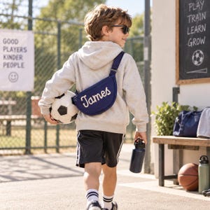May include: A navy blue crescent-shaped crossbody bag featuring the name James embroidered in white script. The bag is worn over a light gray hooded sweatshirt, paired with black athletic shorts, white socks, and a soccer ball held under one arm.