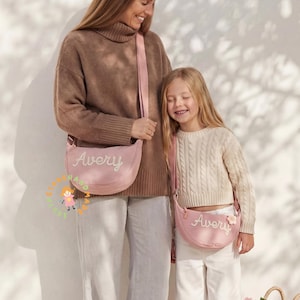 May include: Two matching light pink crescent-shaped crossbody bags featuring the name Avery embroidered in white script. Each bag has a long adjustable shoulder strap and a small flower-shaped charm attached to the side of the smaller bag.