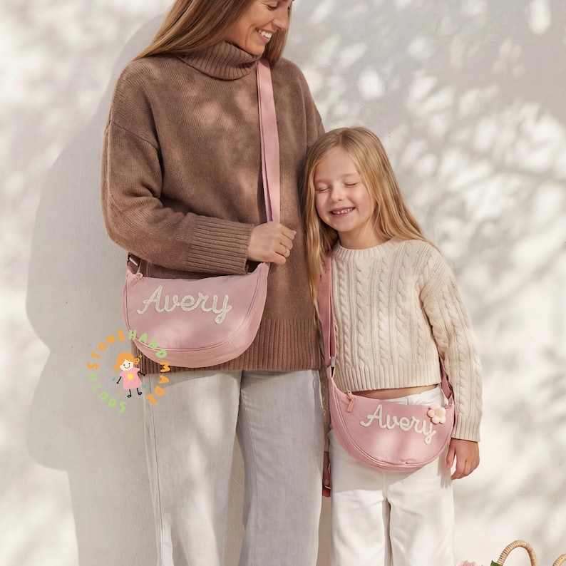May include: Two matching light pink crescent-shaped crossbody bags featuring the name Avery embroidered in white script. Each bag has a long adjustable shoulder strap and a small flower-shaped charm attached to the side of the smaller bag.