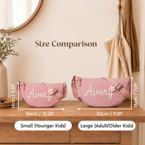 May include: Two pink crescent-shaped crossbody bags with the name Avery embroidered in white and a small flower patch. The small bag measures 10.24 inches by 5.51 inches, and the large bag measures 11.81 inches by 7.09 inches. Text reads Size Comparison.