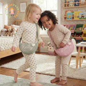 Könnte beinhalten: Zwei Kinder in passenden beigefarbenen Polka-Dot-Pyjamas mit personalisierten Umhängetaschen. Eine Tasche ist salbeigrün mit dem Namen "Reily", die andere rosa mit "Avery". Beide Taschen haben eine weiße Blumenverzierung.