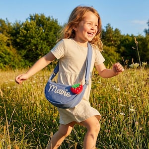 May include: A blue crossbody bag featuring the name Mavie embroidered in white script and a small red strawberry applique. The bag has an adjustable shoulder strap and is worn across the torso over a light beige short-sleeved dress.