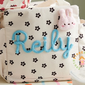 May include: A cream-colored tote bag with a floral pattern and the name "Reily" in blue rope lettering. A pink bunny applique is attached to the bag. The bag has a corduroy texture and a small pocket on the side.