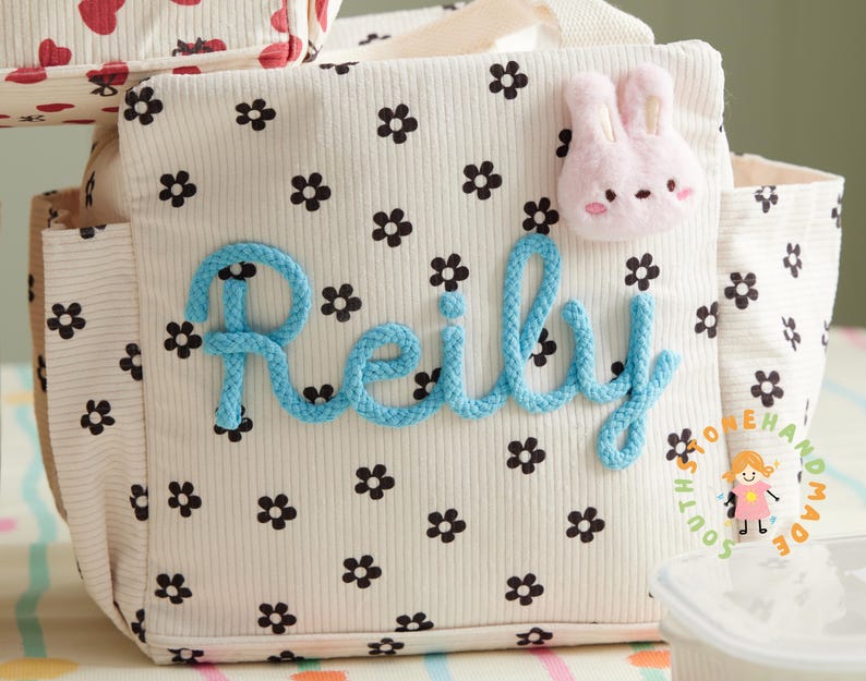 May include: A cream-colored tote bag with a floral pattern and the name "Reily" in blue rope lettering. A pink bunny applique is attached to the bag. The bag has a corduroy texture and a small pocket on the side.