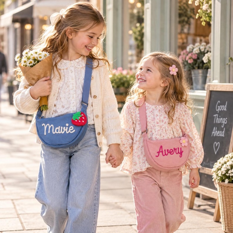 May include: Two crescent-shaped crossbody bags with adjustable straps. The blue bag features the embroidered name Mavie and a strawberry charm. The pink bag features the embroidered name Avery and a flower charm. Both bags have a zipper closure.