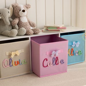 May include: Three fabric storage cubes in beige, pink, and blue, each with a bow and a name in colorful script: Olivia, Callie, and Alice. Plush elephant and dog toys sit above the cubes. A stack of books is also visible.
