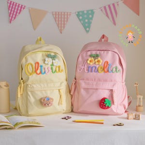 May include: Two personalized backpacks, one yellow and one pink, sit side-by-side. The yellow backpack has the name "Olivia" embroidered in rainbow thread, with floral and bear accents. The pink backpack features the name "Amelia" and a strawberry.