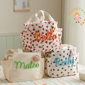 Peut inclure: Trois sacs à lunch personnalisés. Un sac crème avec le nom "Mateo" en vert. Un autre sac blanc avec des cœurs rouges et le nom "Bella" en orange. Le troisième sac est blanc avec des fleurs noires et le nom "Reilly" en bleu.