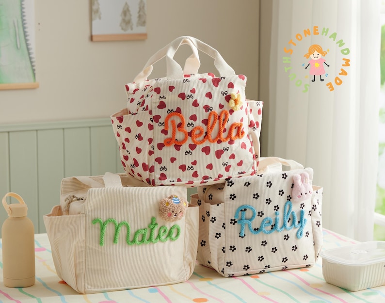 May include: Three personalized lunch bags. One bag is cream-colored with the name "Mateo" in green. Another bag is white with red hearts and the name "Bella" in orange. The third bag is white with black flowers and the name "Reilly" in blue.
