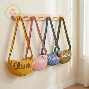 May include: Four crescent-shaped canvas crossbody bags hang from a light wood wall rack. The bags are mustard yellow, dusty pink, blue, and olive green, each embroidered with a name: Oliver, Avery, Mavie, and Charlie, with small floral and strawberry accents.