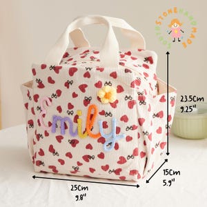 May include: Cream-colored corduroy tote bag with a heart and bow pattern, measuring 9.8" long, 5.9" wide, and 9.25" high. Features the name "Miley" in colorful yarn letters and a yellow flower. Includes a top handle and side pockets.