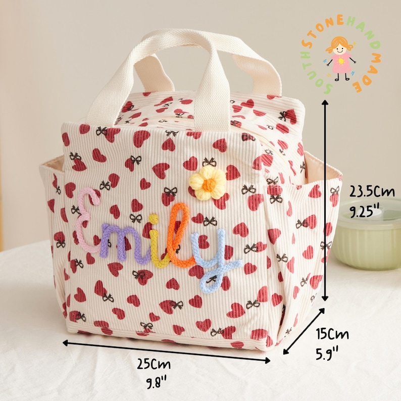 May include: Cream-colored corduroy tote bag with a heart and bow pattern, measuring 9.8" long, 5.9" wide, and 9.25" high. Features the name "Miley" in colorful yarn letters and a yellow flower. Includes a top handle and side pockets.