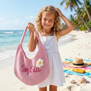 May include: A pink crocheted shoulder bag with the name "Sophia" embroidered on the front, and a white daisy applique. The bag is held by a person on a beach. A straw hat, sunscreen, and a colourful towel are on the sand.