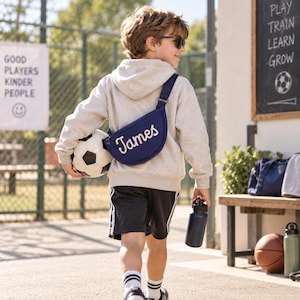 May include: A navy blue crescent-shaped crossbody bag featuring the name James embroidered in white script. The bag is worn over a light gray hooded sweatshirt, paired with black athletic shorts, white socks, and a soccer ball held under one arm.