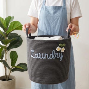 May include: A dark grey rope laundry basket with the word "Laundry" embroidered in light blue. The basket has two handles and is decorated with white and green floral accents. The basket is filled with white fabric.