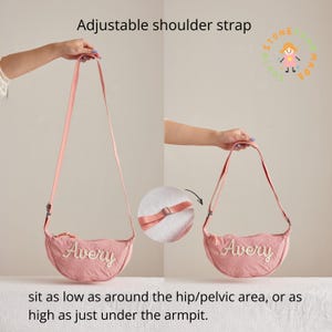 May include: A pink crescent-shaped crossbody bag featuring the name Avery embroidered in white script on the front. The bag includes an adjustable shoulder strap with a metal slider, allowing it to be worn at various lengths from the hip to the armpit.