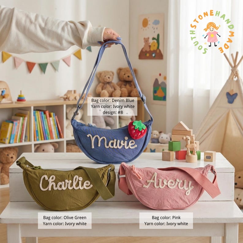 May include: Three crescent-shaped crossbody bags in denim blue, olive green, and pink. Each bag features a personalized name embroidered in ivory white yarn. The denim blue bag includes a small strawberry charm. The bags have adjustable straps.
