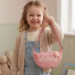 Op de afbeelding: Een roze, gewatteerde crossbodytas met de naam "Avery" in witte letters geborduurd. De tas heeft een bloemenapplicatie en een lange riem. Het kind draagt een lichtroze vest, een spijkerbroek en een crèmekleurig t-shirt.