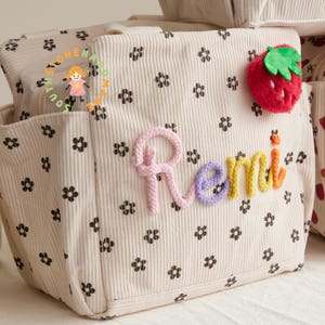 May include: Cream-colored corduroy tote bag with black floral pattern and the name "Remi" in colorful yarn. A red strawberry with green leaves is attached to the side. The bag has multiple pockets and a zipper closure.