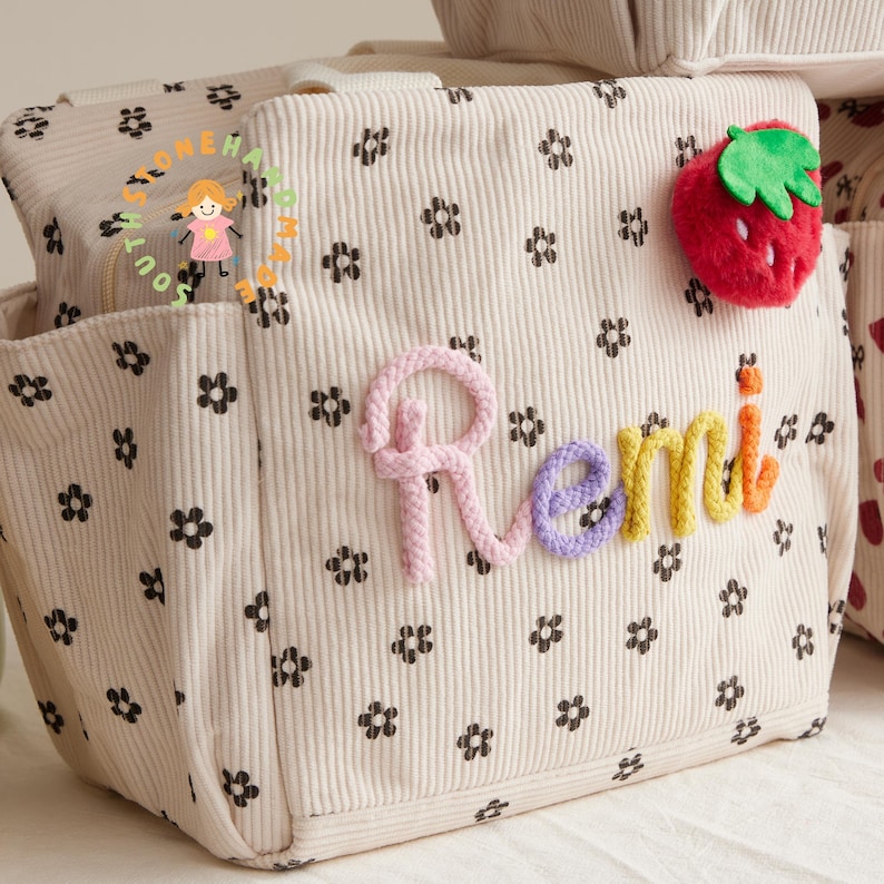 May include: Cream-colored corduroy tote bag with black floral pattern and the name "Remi" in colorful yarn. A red strawberry with green leaves is attached to the side. The bag has multiple pockets and a zipper closure.