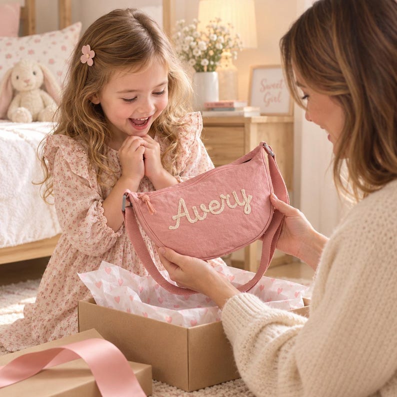 May include: A dusty pink crescent-shaped crossbody bag featuring the name Avery embroidered in cream-colored thread on the front. The bag has a matching pink adjustable strap and a zipper closure, presented as a gift in a cardboard box.