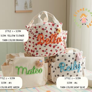 May include: Three personalized lunch bags with different designs. One bag is white with red hearts and the name "Bella" in orange yarn. Another is beige with "Mateo" in green yarn. The third is white with black flowers and "Reilly" in blue yarn.