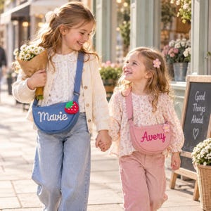 May include: Two crescent-shaped crossbody bags with adjustable straps. The blue bag features the embroidered name Mavie and a strawberry charm. The pink bag features the embroidered name Avery and a flower charm. Both bags have a zipper closure.