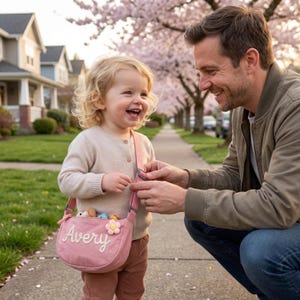 May include: A dusty pink crossbody bag featuring the name Avery embroidered in white script lettering on the front, accented with a small, light-colored flower applique, and equipped with an adjustable strap for carrying.