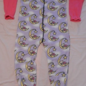 May include: A one-piece footed pajama suit featuring long pink sleeves and a lavender body with a repeating pattern of teddy bears sleeping on yellow crescent moons and stars. The garment includes a full-length front zipper closure and blue soles.