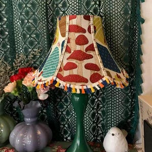 Op de afbeelding: Een groene tafellamp met een lampenkap met een paddenstoelmotief. De lampenkap heeft een witte achtergrond met rode en gele paddenstoelen. De lamp heeft een groene voet en een witte kwastversiering.