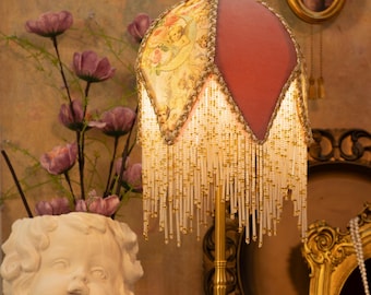 May include: A vintage-style table lamp with a gold-colored base and a decorative lampshade. The lampshade features a floral pattern and a red velvet section, adorned with beaded fringe. The lamp is placed near a cherub statue and flowers.