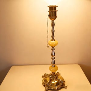 Może przedstawiać: Zabytkowa mosiężna lampa stołowa z dekoracyjną podstawą i łańcuszkiem. Lampa posiada dwie żółte szklane kule i szczegółowy, ozdobny design. Lampa stoi na jasnej powierzchni.