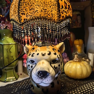 May include: A table lamp with a leopard print lampshade and a ceramic cheetah head base. The lampshade has black beads around the bottom edge. The lamp is sitting on a black lace tablecloth.