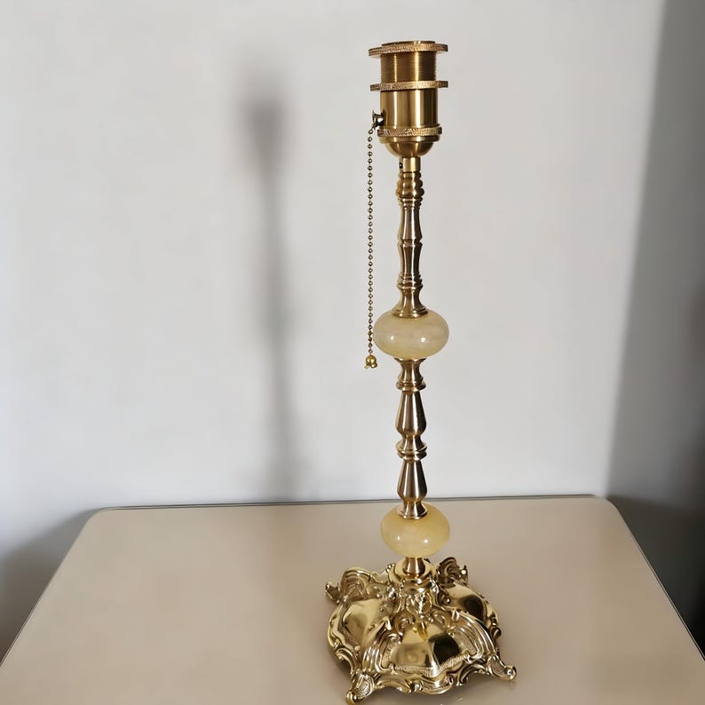 May include: A vintage brass table lamp with a decorative base and a central column featuring two alabaster-like orbs. The lamp has a pull chain and a standard lamp socket. The base is ornate and detailed.