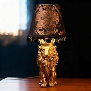 Op de afbeelding: Goudkleurige tafellamp in de vorm van een leeuw met een kroon. De zwarte lampenkap heeft een gouden bloemenpatroon en donkergroene pompom-afwerking. De lamp staat op een houten oppervlak. Hoogte ongeveer 50 cm.
