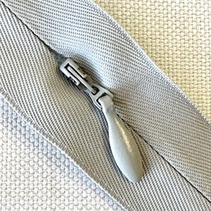 May include: Close-up of a light gray zipper with a matching fabric background. The zipper pull is a rounded, metallic gray. The fabric has a woven texture, and the zipper is partially open, revealing the seam.