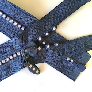 May include: Navy blue zipper with decorative rhinestones. The zipper is crossed over itself, showing the teeth and pull tab. The rhinestones are set in a dark metal. The zipper is on a white background.