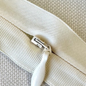 May include: Close-up of an off-white zipper with a matching pull tab. The zipper tape has a woven texture, and the pull tab is a teardrop shape. The zipper is partially open, revealing the teeth.