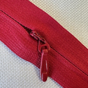 May include: Close-up of a red zipper on a textured fabric. The zipper pull is a matching red color. The fabric is a deep red, and the background is a light beige.