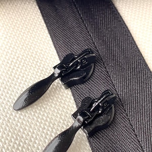 May include: Close-up of two black zipper pulls on a gray fabric. The pulls have a teardrop shape and are set against a textured, off-white background. The zipper teeth are visible at the top of the image.