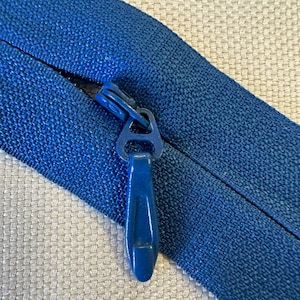 May include: A close-up of a blue zipper with a blue plastic pull tab. The zipper is closed and the pull tab is in the up position.