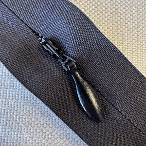 May include: Close-up of a black zipper with a teardrop-shaped pull. The zipper is on a dark navy blue fabric, with a contrasting light gray woven fabric in the background. The zipper's metal components are also black.