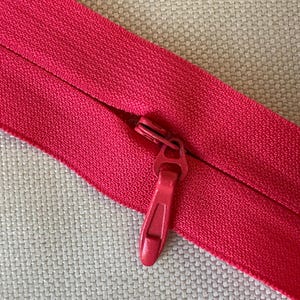 May include: Close-up of a bright red zipper on a textured fabric background. The zipper is partially open, revealing the teeth. The pull tab is also red. This item is suitable for sewing projects.