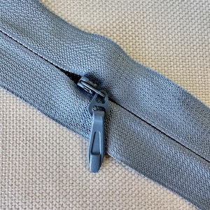 May include: Close-up of a blue zipper with a matching pull tab. The zipper is partially open, revealing the teeth. The fabric is a textured, light blue material. The background is a neutral, woven fabric.
