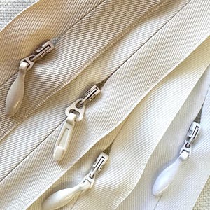 May include: Close-up of several beige zippers with matching pulls. The zippers are arranged diagonally, showcasing the fabric texture and the metal and plastic components. The pulls have a teardrop shape.