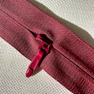 May include: Close-up of a burgundy zipper on a textured fabric background. The zipper pull is a matching burgundy color. The zipper is partially open, revealing the teeth and the fabric's weave.