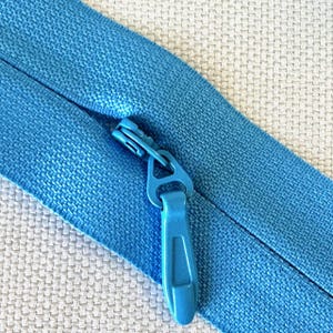 May include: Close-up of a bright blue zipper with a matching pull tab. The zipper is partially open, revealing the teeth and the fabric it is sewn into. The fabric has a textured appearance.