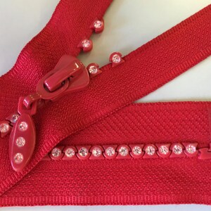 Puede incluir: Primer plano de una cremallera roja con un tirador a juego y pedrería decorativa. La cinta de la cremallera es de color rojo vibrante y la pedrería añade un toque de brillo. La cremallera está diseñada para proyectos de costura.